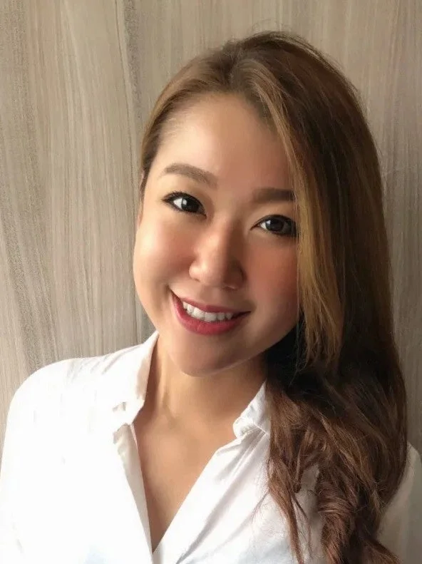 白いシャツを着た、茶色の長い髪の女性が笑顔でカメラを見ているポートレート写真です。明るい背景の前で、親しみやすい表情をしています。