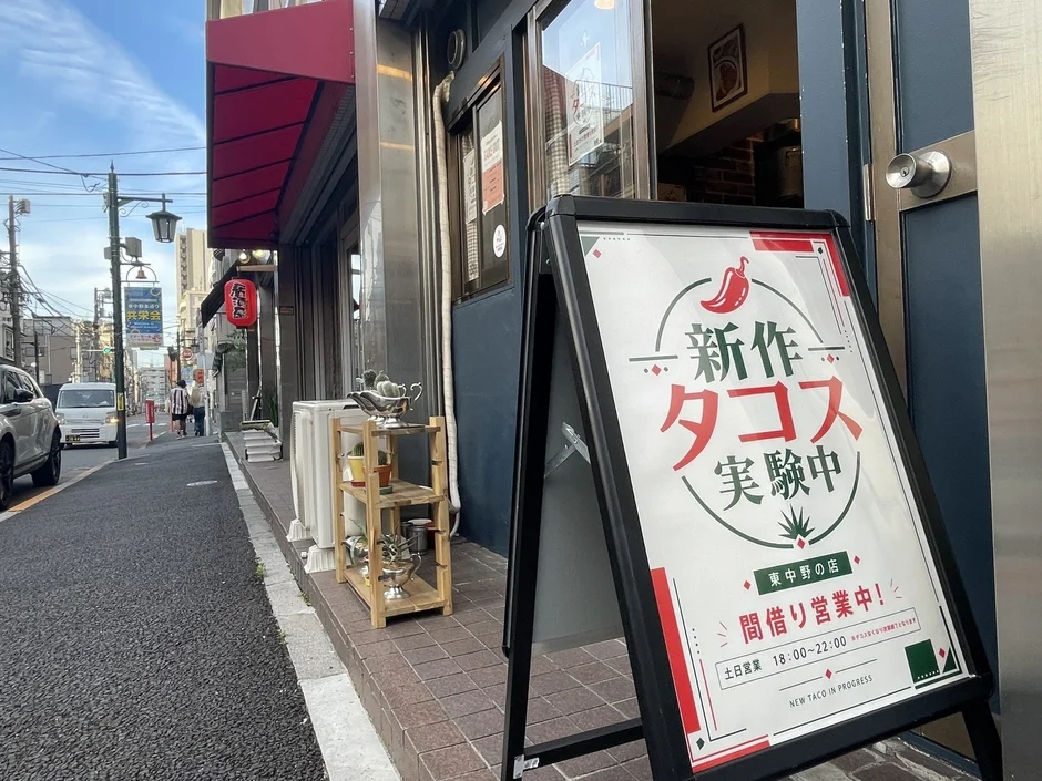 日本の街角にある店舗の前で、新作タコスの間借り営業を告知するA型看板が置かれている様子を捉えた画像です。営業は土日18時から22時と記載されています。