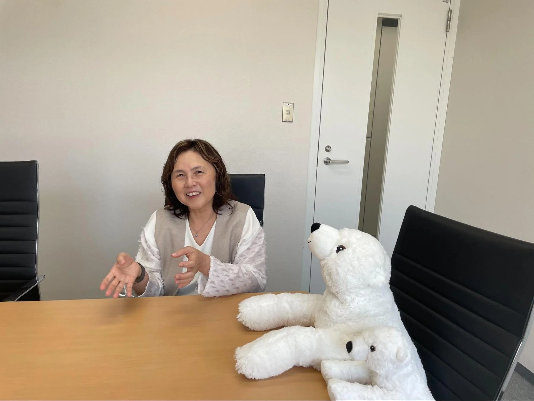会議室のような場所で、笑顔の女性が木製のデスクに座り、身振り手振りを交えながら話しています。隣には親子のシロクマのぬいぐるみが置かれています。