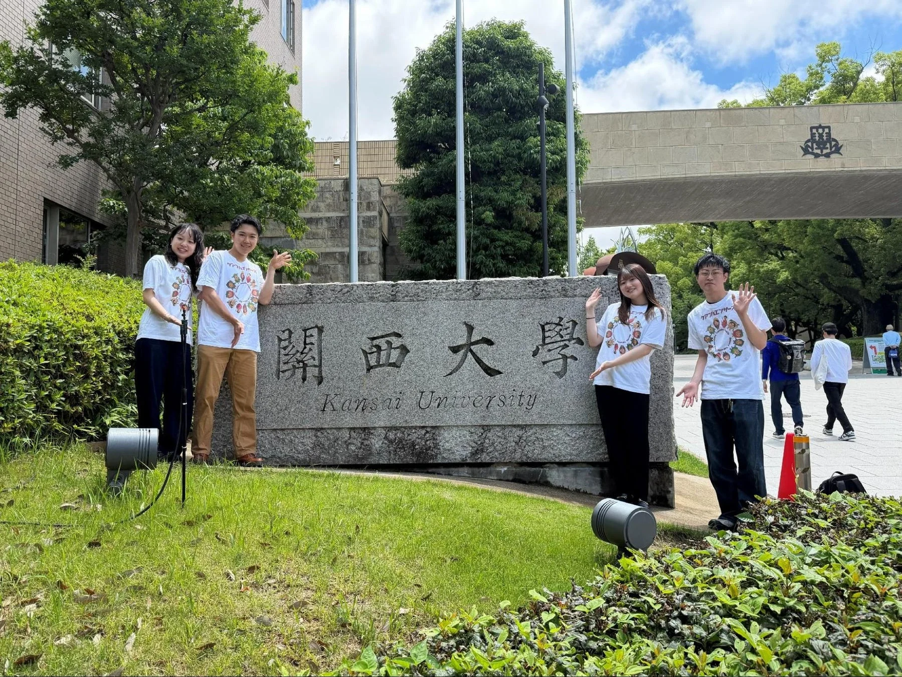 関西大学の石碑前で笑顔でポーズをとる4人の若者