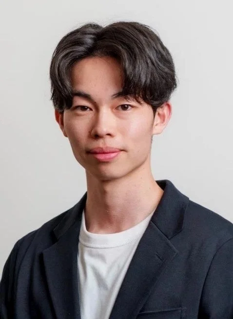 明るい背景の前でカメラを見つめる若いアジア人男性のポートレート。黒いジャケットと白いTシャツを着用し、落ち着いた表情をしている。