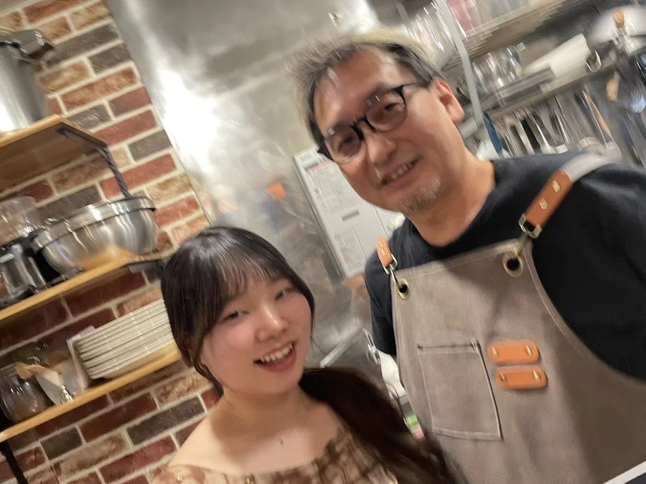 厨房のような場所で、エプロンを着用した男性と女性が笑顔でカメラに写っています。楽しそうな雰囲気のセルフィーです。