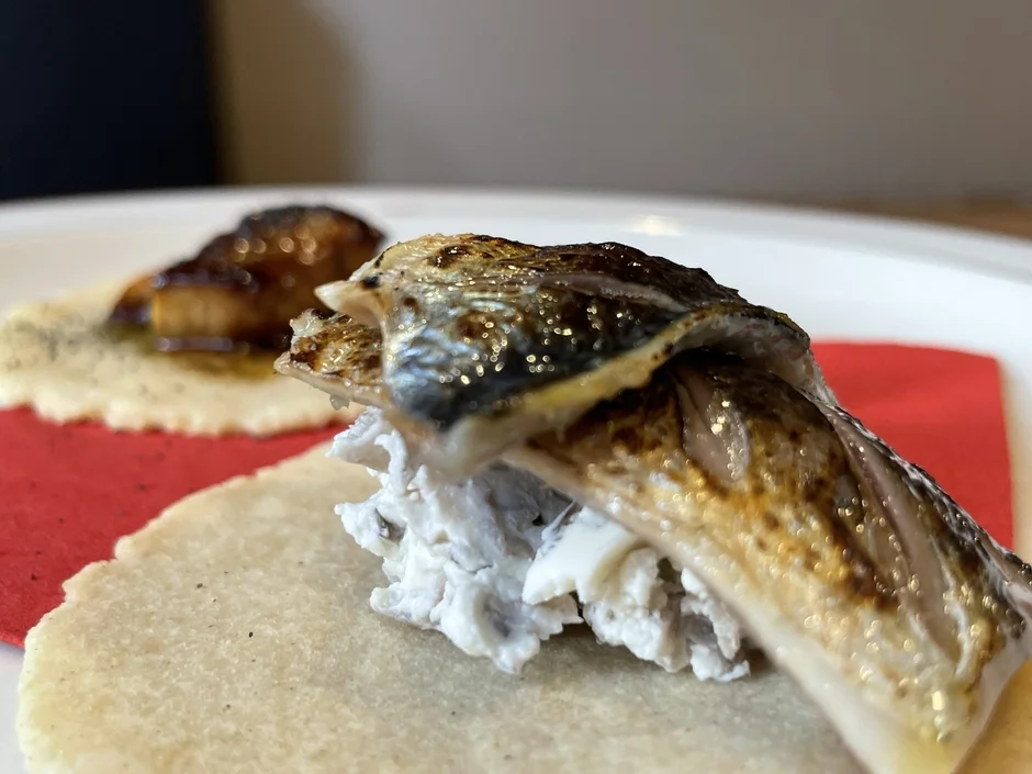 香ばしく焼き上げられた魚が、白いクリームとフラットブレッドの上に美しく盛り付けられた一品。奥には別の料理もぼやけて写っており、洗練されたレストランでの食事風景を思わせる。