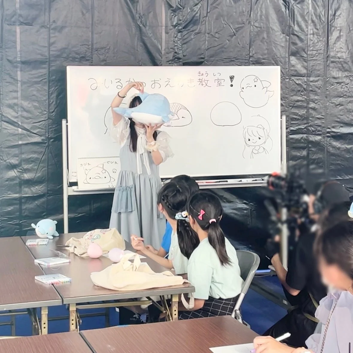 女性がイルカのぬいぐるみを使って、子供たちに絵の描き方を教えている様子