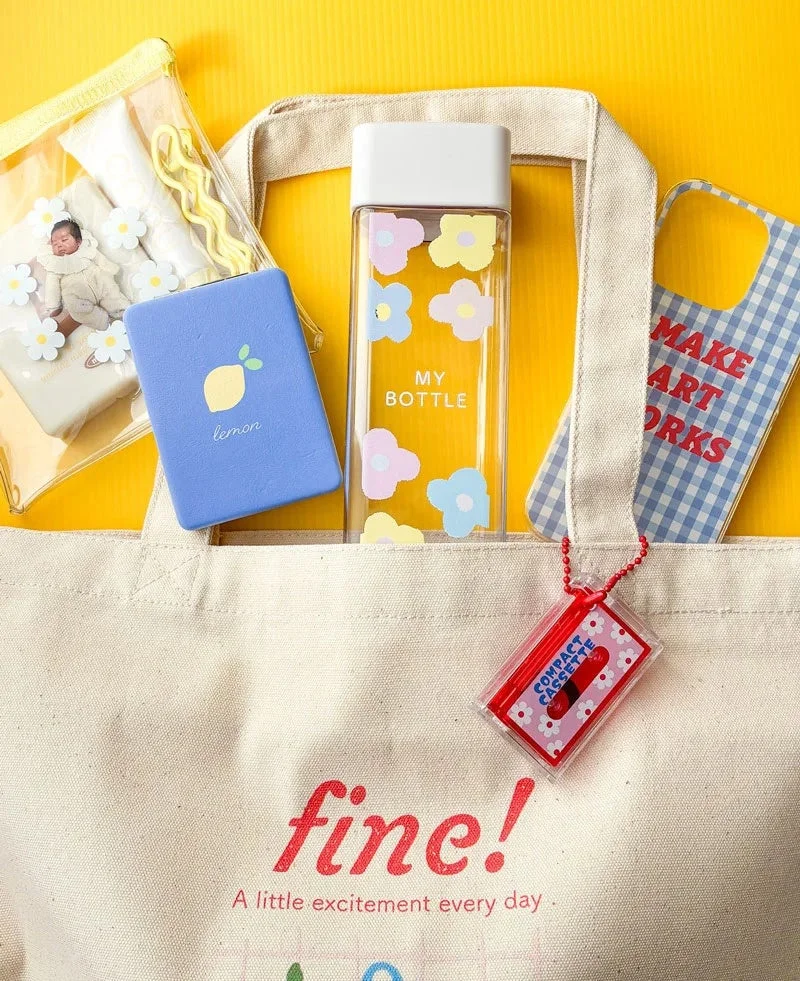 黄色い背景の上に、ベージュのトートバッグと、花柄の水筒、レモン柄のケース、チェック柄のスマホケースなど、カラフルで可愛らしい日用品や雑貨が並べられています。
