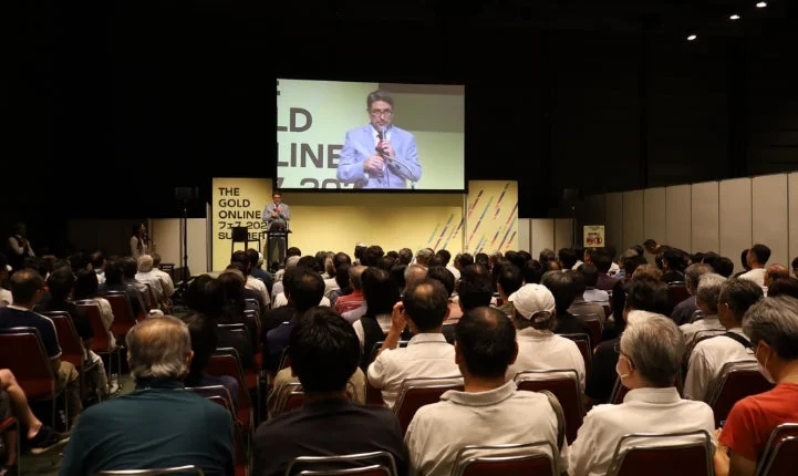 大勢の聴衆が参加する屋内イベントで、ステージ上の講演者がスクリーンに映し出されながらプレゼンテーションを行っています。